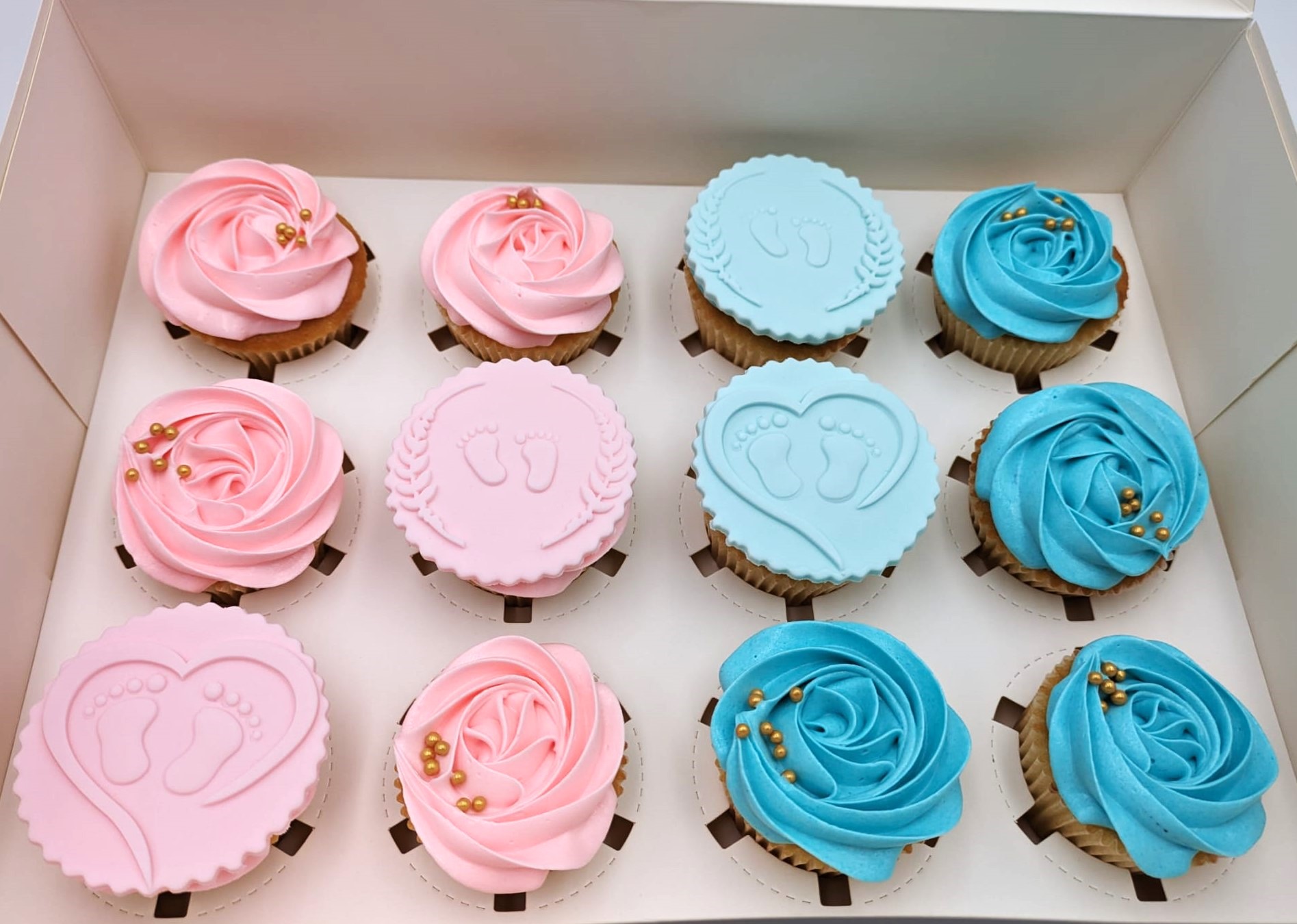 A dozen gender reveal cupcakes in a box, blue and pink colors with tiny feet and swirl decor.